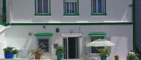 A two-story building with a white facade and green trim. There are three windows with shutters, a door, and a small balcony with a striped umbrella.