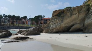 Ein Strand mit großen Felsen, sandigem Ufer und Gebäuden im Hintergrund.