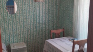 Ein Zimmer mit Streifenmuster-Tapete, ein kleiner Tisch mit weißem Tuch, ein Holzstuhl und ein weißes Regal.