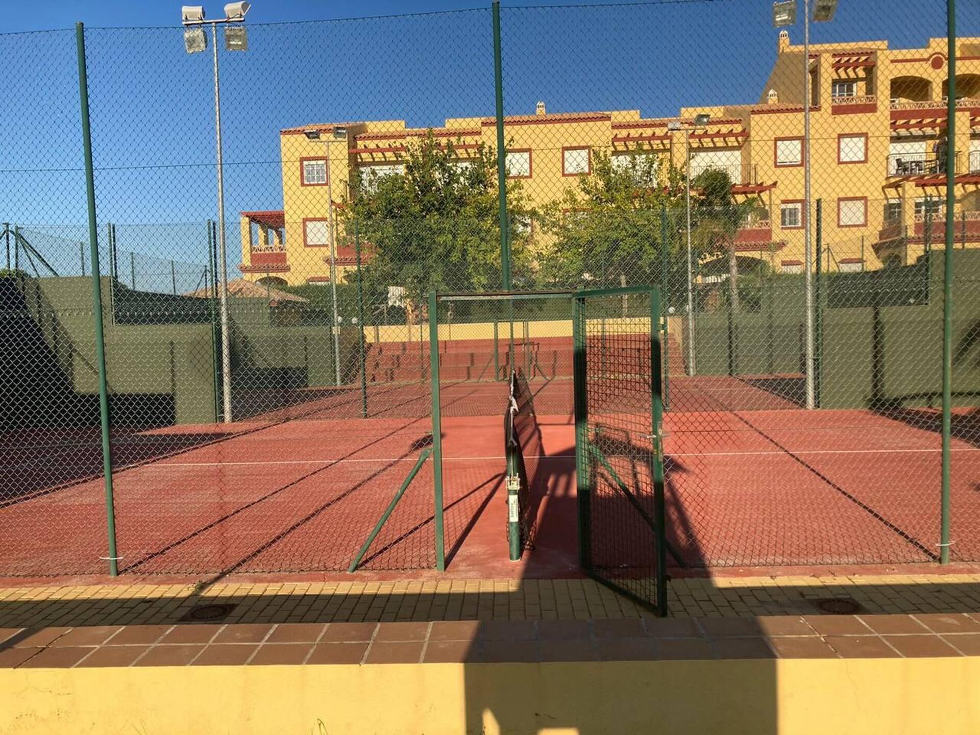 En tennisbane med hegn af kædetråd, omgivet af bygninger.