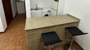Eine kleine Küchenzeile mit einer Mikrowelle, einer Waschmaschine und einem Spülbecken. Zwei Barhocker mit schwarzen Sitzflächen sind vorhanden.