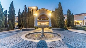 Μια αυλή με ένα κεντρικό σιντριβάνι, που περιβάλλεται από πλακόστρωτα μονοπάτια και ψηλά δέντρα.