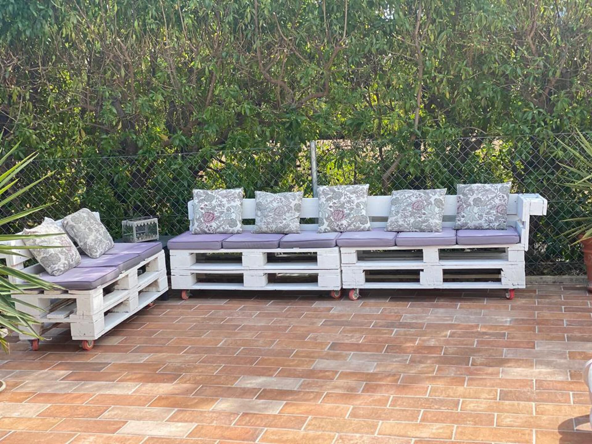 A white wooden bench with cushions and a small table, set on a brick patio with a green hedge in the background.