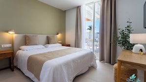 Ein Hotelzimmer mit einem großen Bett, einem Nachttisch aus Holz, einem kleinen Tisch mit einer Lampe und Blick ins Freie durch eine Schiebetür.
