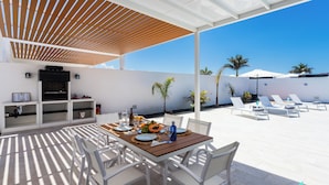 Eine Terrasse mit Esstisch für vier Personen, umgeben von weißen Stühlen, unter einer Holzpergola.