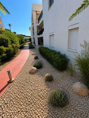 Un chemin menant à un bâtiment dont l’entrée est ornée d’un tapis rouge, et qui est entouré de gravier, de rochers et de diverses plantes.