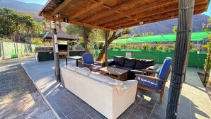 Eine Terrasse im Freien mit überdachter Sitzgelegenheit, einem Grill und Blick auf ein grünes Feld und Berge.