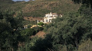 Un grand bâtiment d’un seul étage, doté d’une façade blanche et d’un toit brun, entouré d’une végétation luxuriante et avec des collines en arrière-plan.
