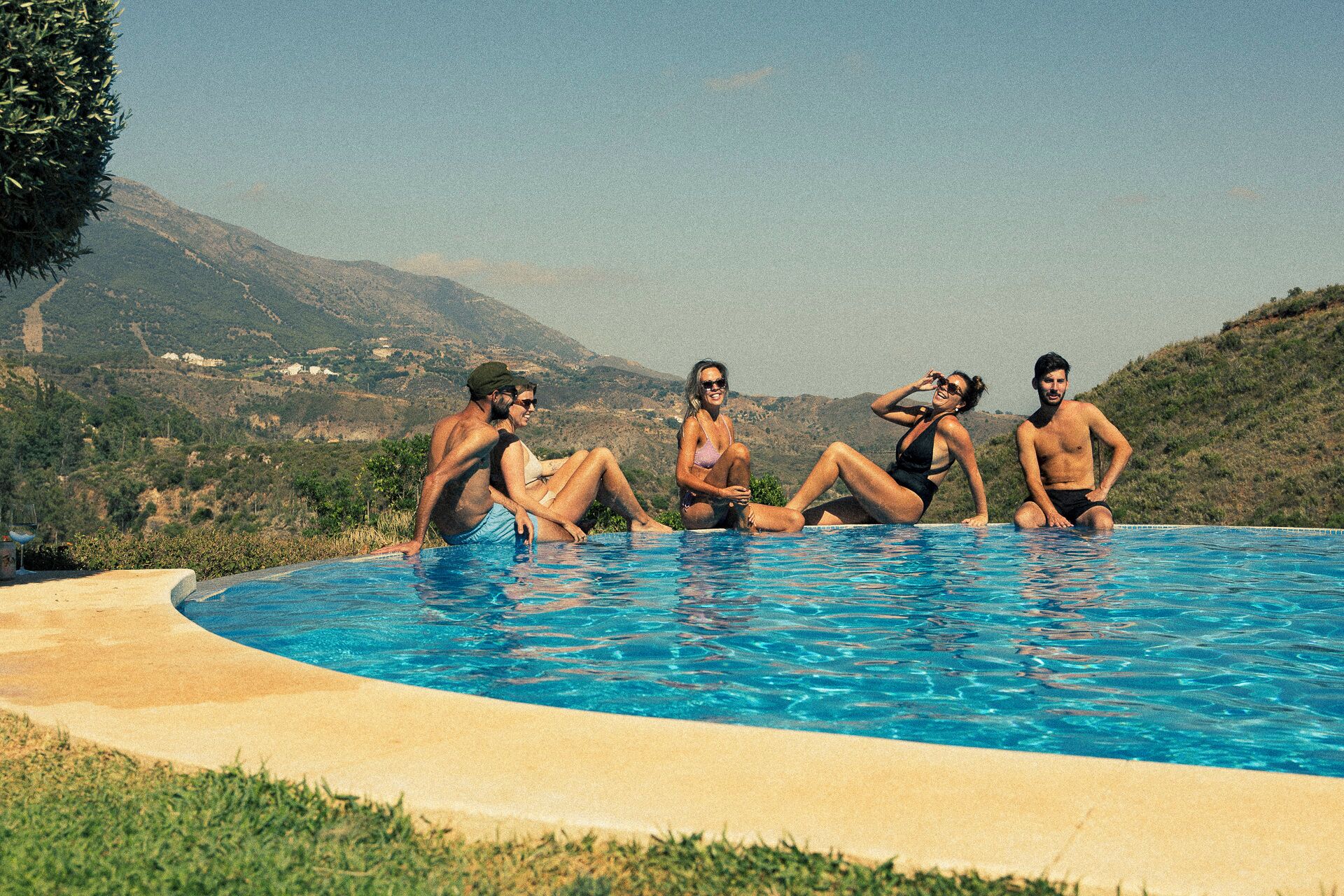 A group of people sitting by a swimming pool with a mountainous landscape in the background.