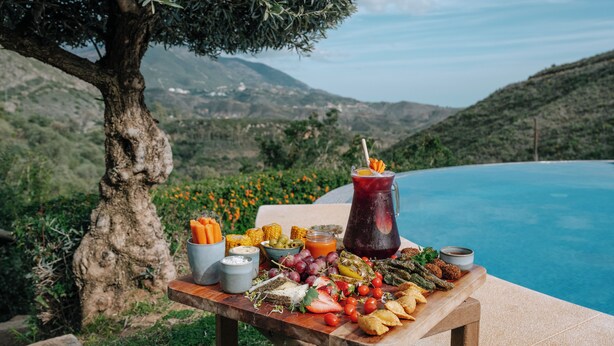 Une table dressée avec une variété de plats et de boissons, le tout mis en valeur par un décor pittoresque composé d’une piscine et de montagnes.
