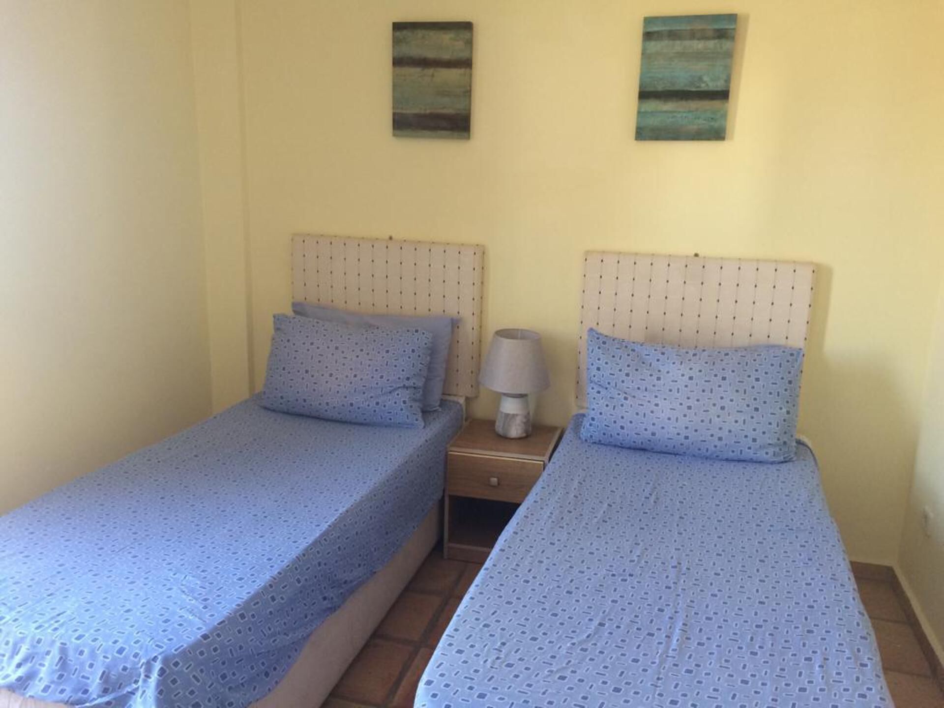 Une chambre avec deux lits simples, chacun doté d’une tête de lit et d’un oreiller. Une table de chevet avec une lampe se trouve entre les deux lits.