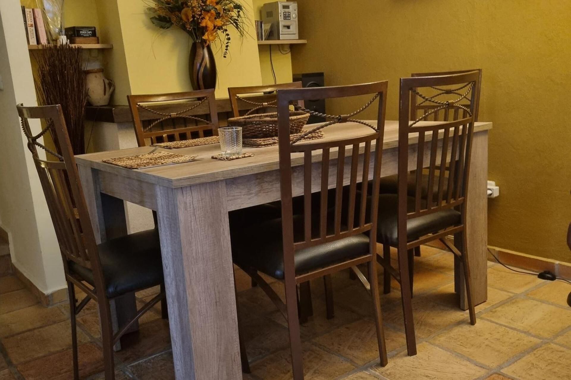 A dining table with four wooden chairs, a vase with flowers, and a radio on a shelf.