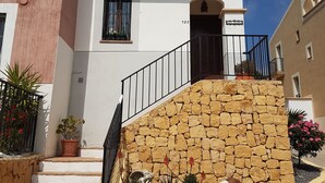 Un bâtiment à deux étages, avec un escalier en pierre menant à l’entrée, un balcon agrémenté de plantes en pot et un ciel bleu clair en arrière-plan.