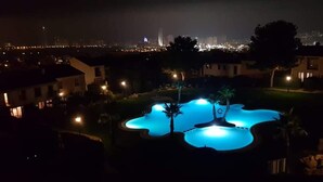 Vue nocturne d’un quartier résidentiel avec une piscine illuminée de lumières bleues, entourée de maisons et, au loin, de l’horizon urbain de la ville.