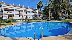 A swimming pool with a clear blue water, surrounded by a white building with balconies, palm trees, and a grassy area. - Casa Buganvilla 11 at Lauro Golf (Alhaurin de la Torre)