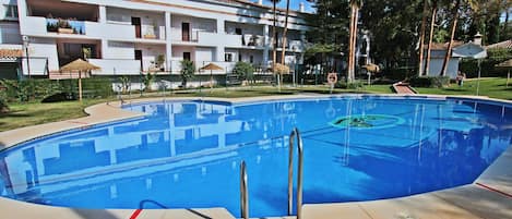 Una piscina con agua cristalina azul, rodeada por un edificio blanco con balcones, palmeras y un área verde.
