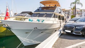 Een witte boot afgemeerd in een jachthaven, met een overdekte cabine en een rode vlag aan de achterkant.