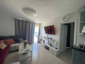 A living room with a sofa, a coffee table, a TV, a clock, and a kitchen area with cabinets. - Apartment - Sleeps 4 - Jacuzzi - Terrace - Pool (Adeje)