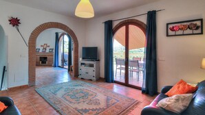 A living room with a fireplace, a television, a sofa, and a large rug. There are two doors leading to an outdoor area. - Casa Redonda a Tranquil Spacious Apartment (Còmpeta)