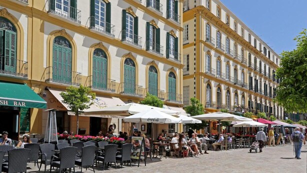 A bustling outdoor café area with outdoor seating and umbrellas, in front of a historic building with multiple balconies and green shutters. - Malaga Cityheart Stay Bright Modern City (Málaga)