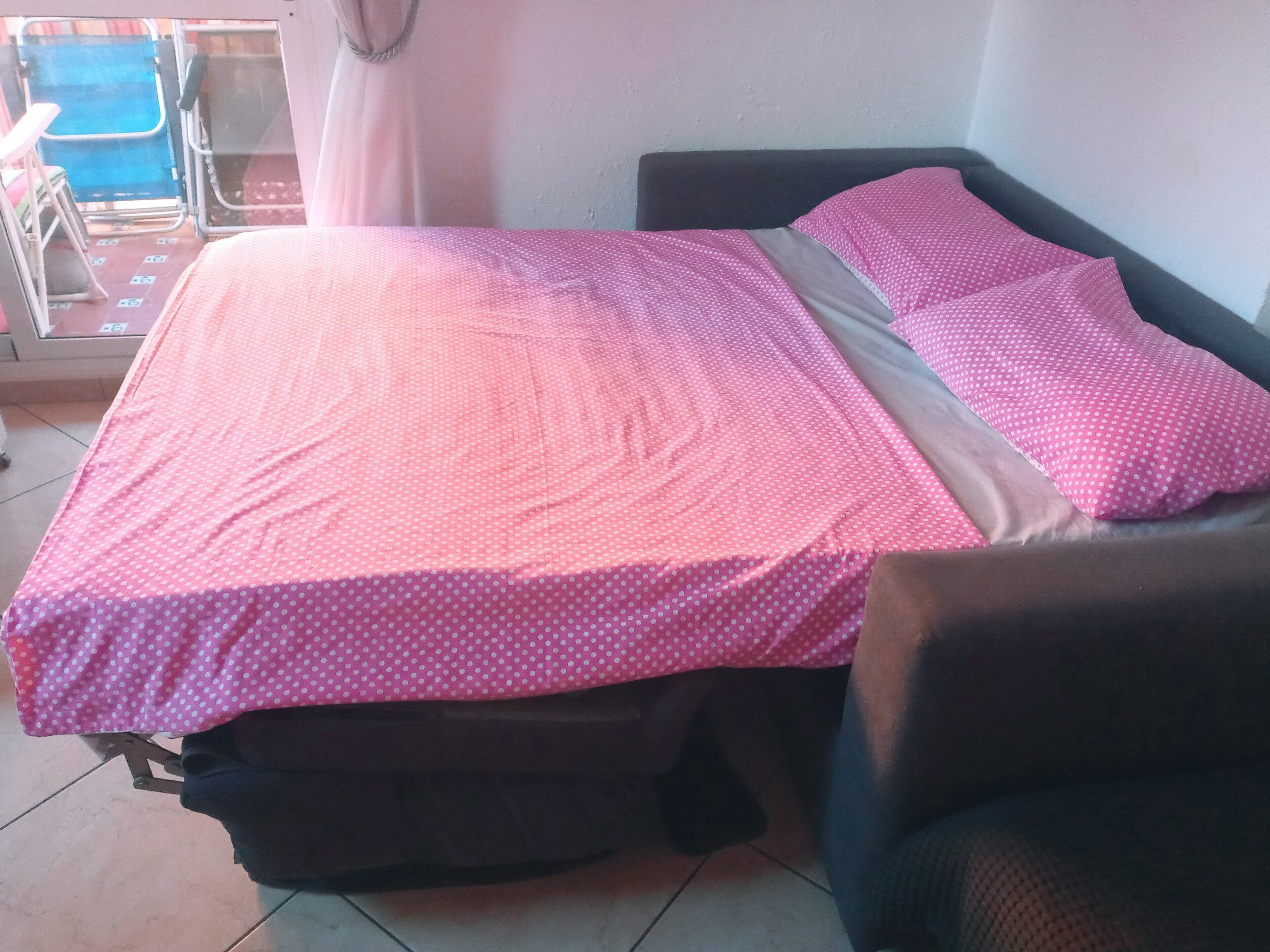 Una cama con una colcha rosa de lunares y almohadas a juego, un cabecero oscuro y una silla a la derecha.