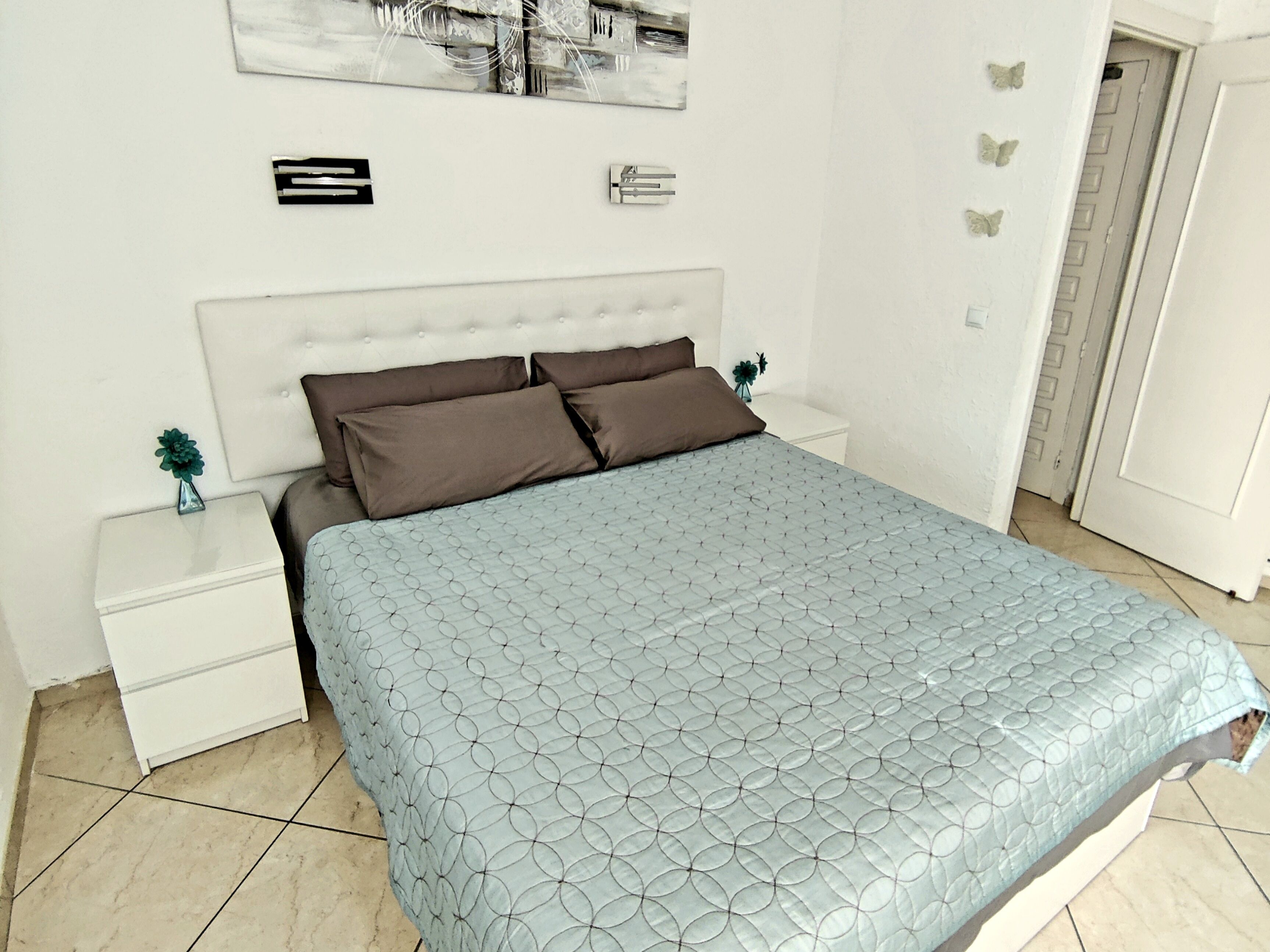 Una cama bien hecha con una colcha estampada, cabecera blanca y dos mesitas de noche con pequeños adornos.