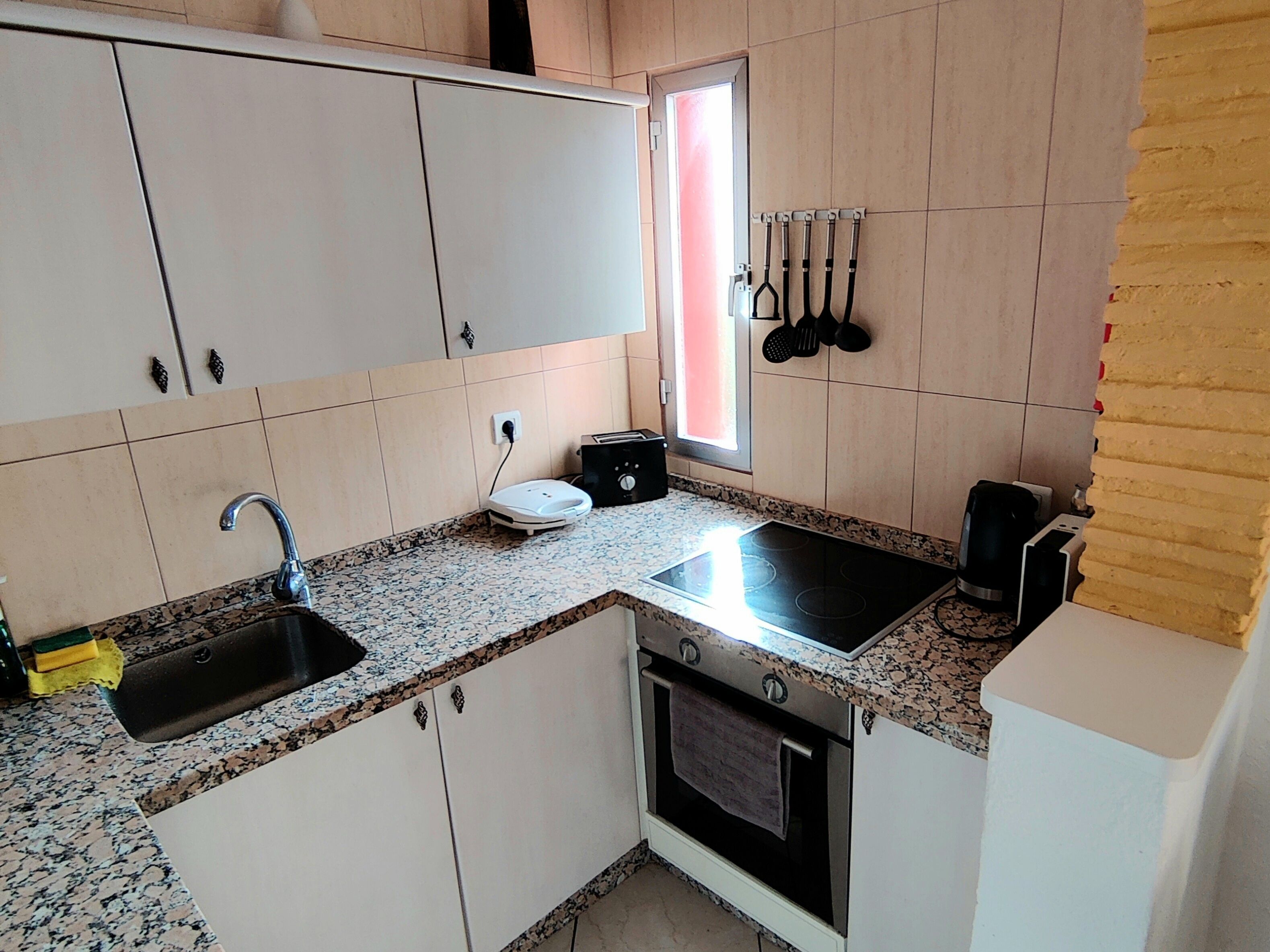 Une cuisine de taille réduite avec des meubles blancs, un plan de travail en granit, un four encastré, un évier et une fenêtre agrémentée d’un rideau rouge.