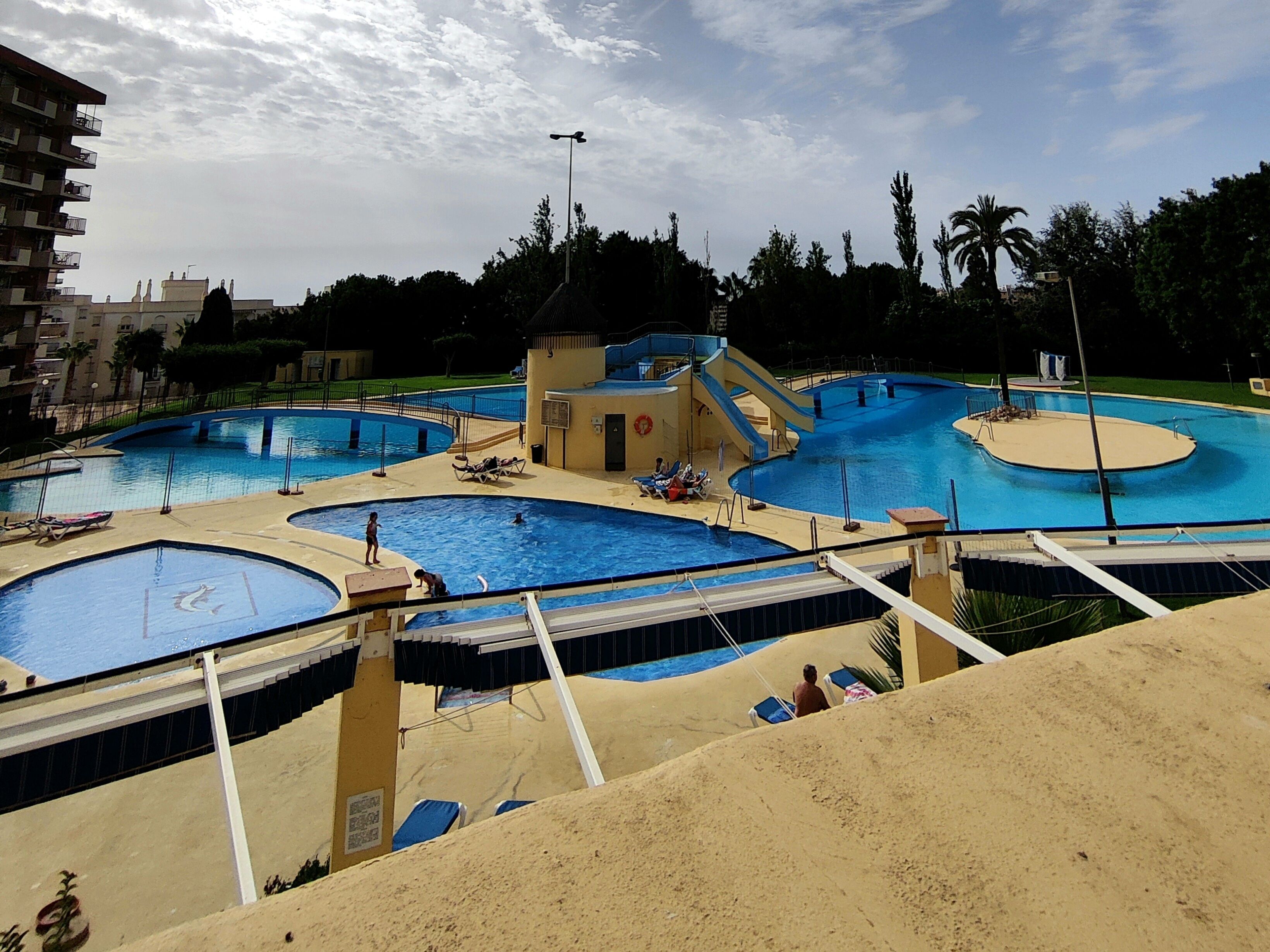 Un grand complexe de piscines extérieures comprenant plusieurs bassins et un petit bâtiment, le tout entouré de palmiers et surplombé par un ciel dégagé.