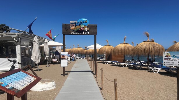A beachside walkway with thatched umbrellas and lounge chairs, a sign indicating 'Despega 1974', and a small kiosk. - La Cala 2 Bedroom Apt With Sea View and Pool View (Mijas)