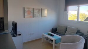 Un salon moderne doté d’un téléviseur à écran plat, d’un canapé, d’une table basse et d’une fenêtre offrant une vue sur des immeubles et des arbres.