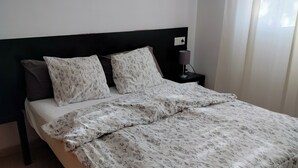 Un lit bien fait, avec une literie à motifs floraux, une tête de lit sombre et une table de chevet surmontée d’une lampe.