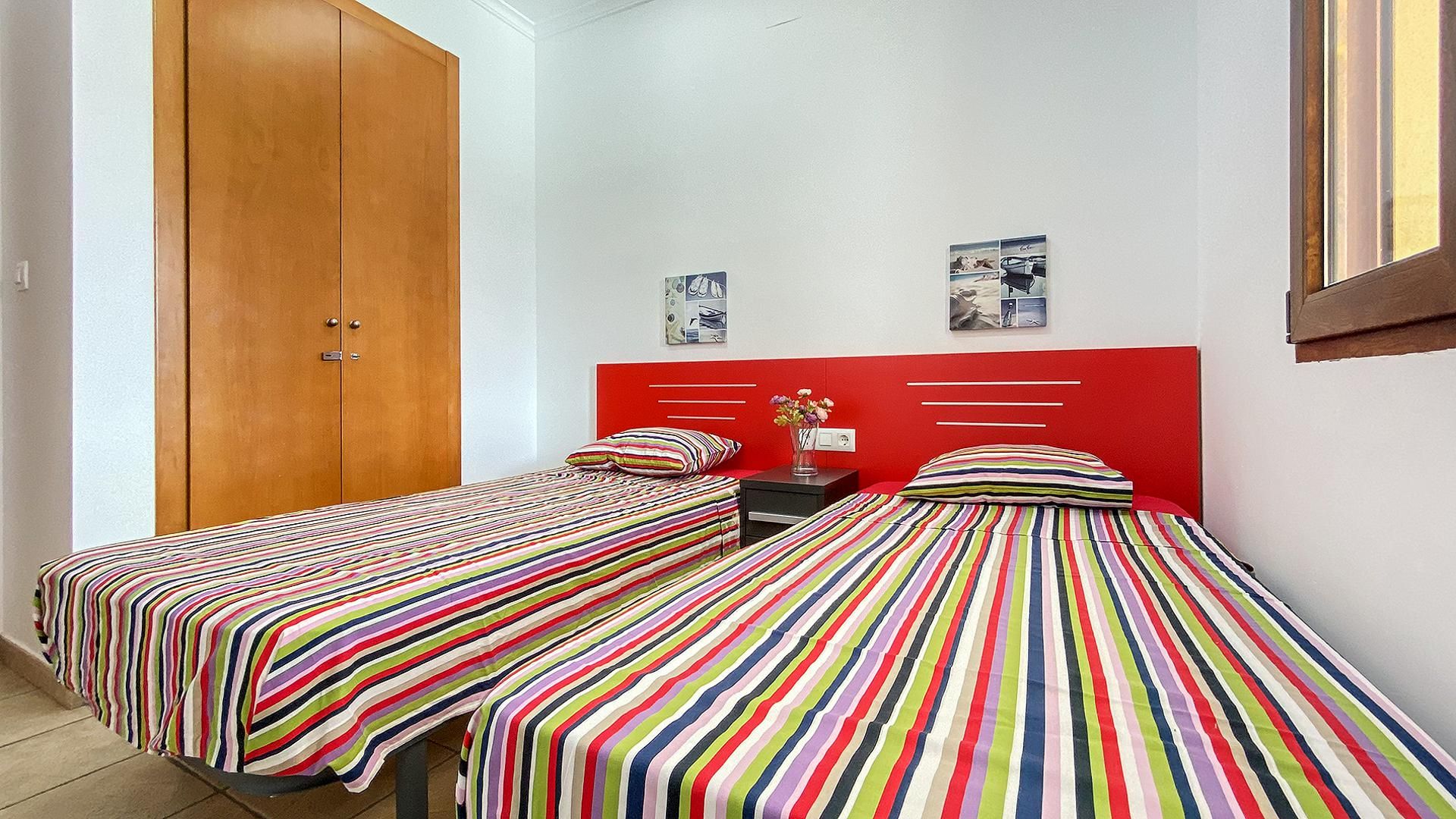 A bedroom with two single beds, a wooden wardrobe, a red headboard, and a small bedside table with a vase.