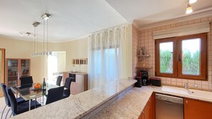 A modern kitchen with a dining area, a dishwasher, and a window with a view of greenery. - Villa El Palmar 2 en Denia (Denia)