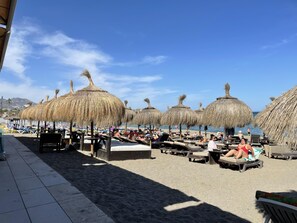 Een strandgebied met rieten parasols, ligbedden en een betegeld pad.