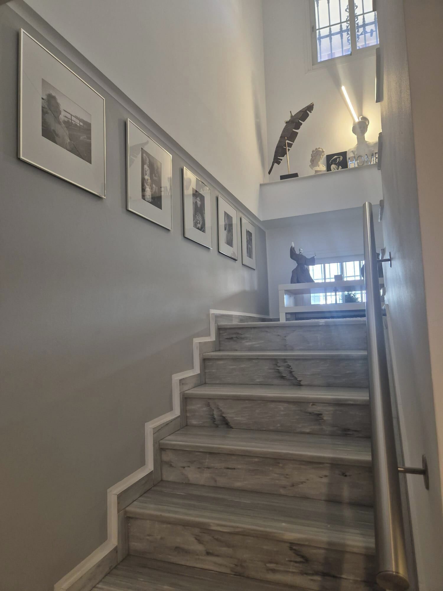 Una escalera de mármol con barandilla, que lleva a un rellano con una estatua y cuadros enmarcados en la pared.