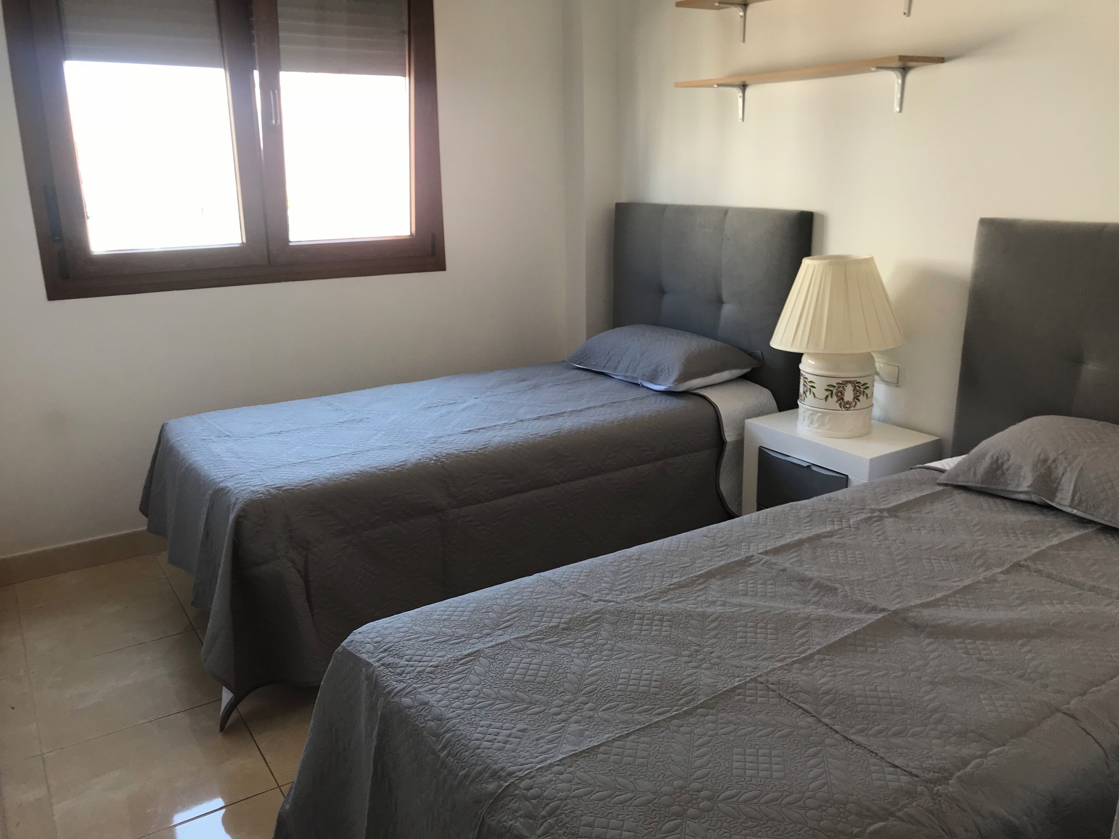 A bedroom with two beds, a nightstand with a lamp, and a window with blinds.