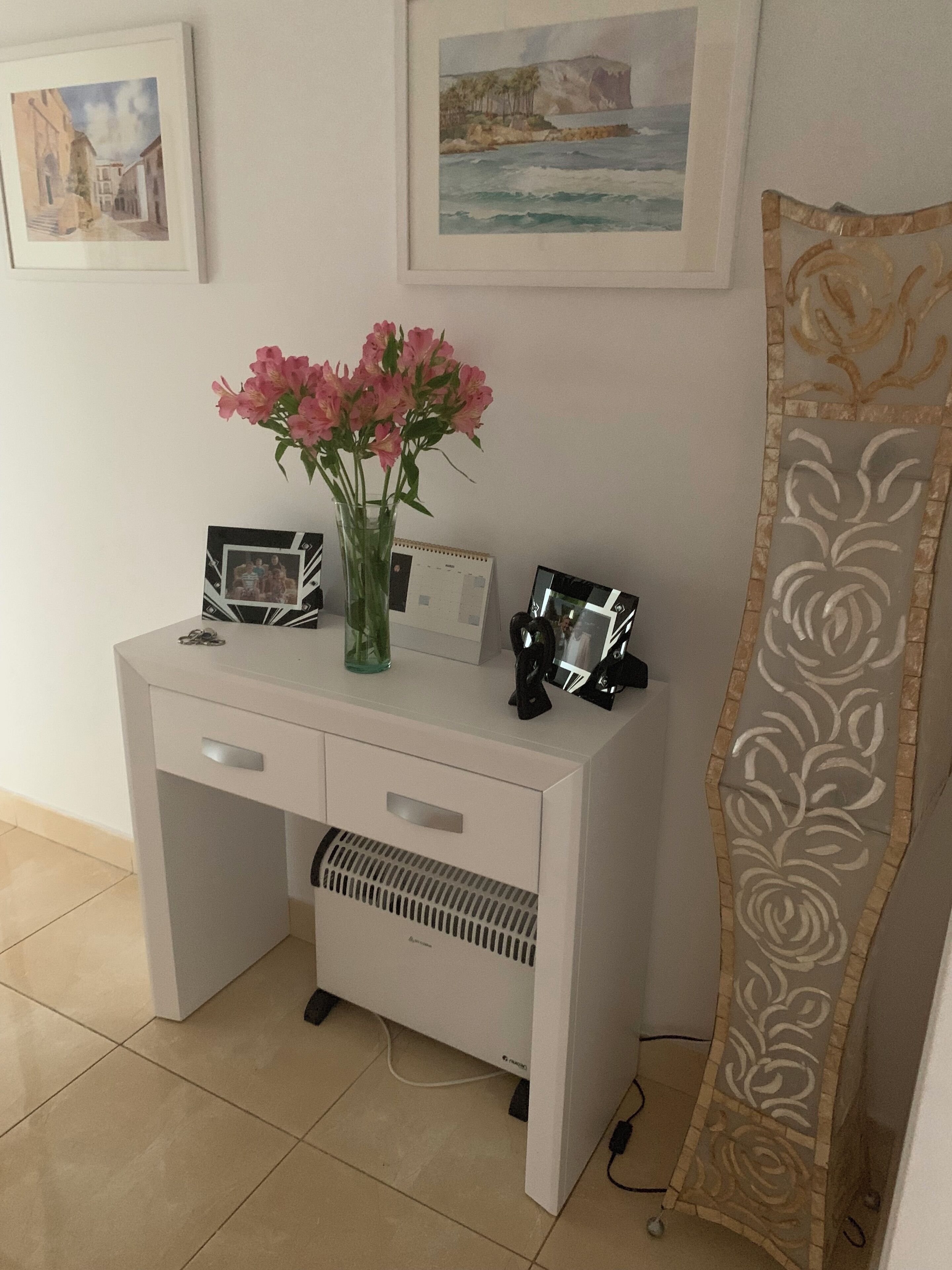 A white desk with a vase of pink flowers, a framed photo, and a calendar.