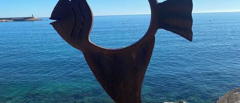 A metal sculpture of a fish in a coastal setting with clear blue water and rocky terrain.