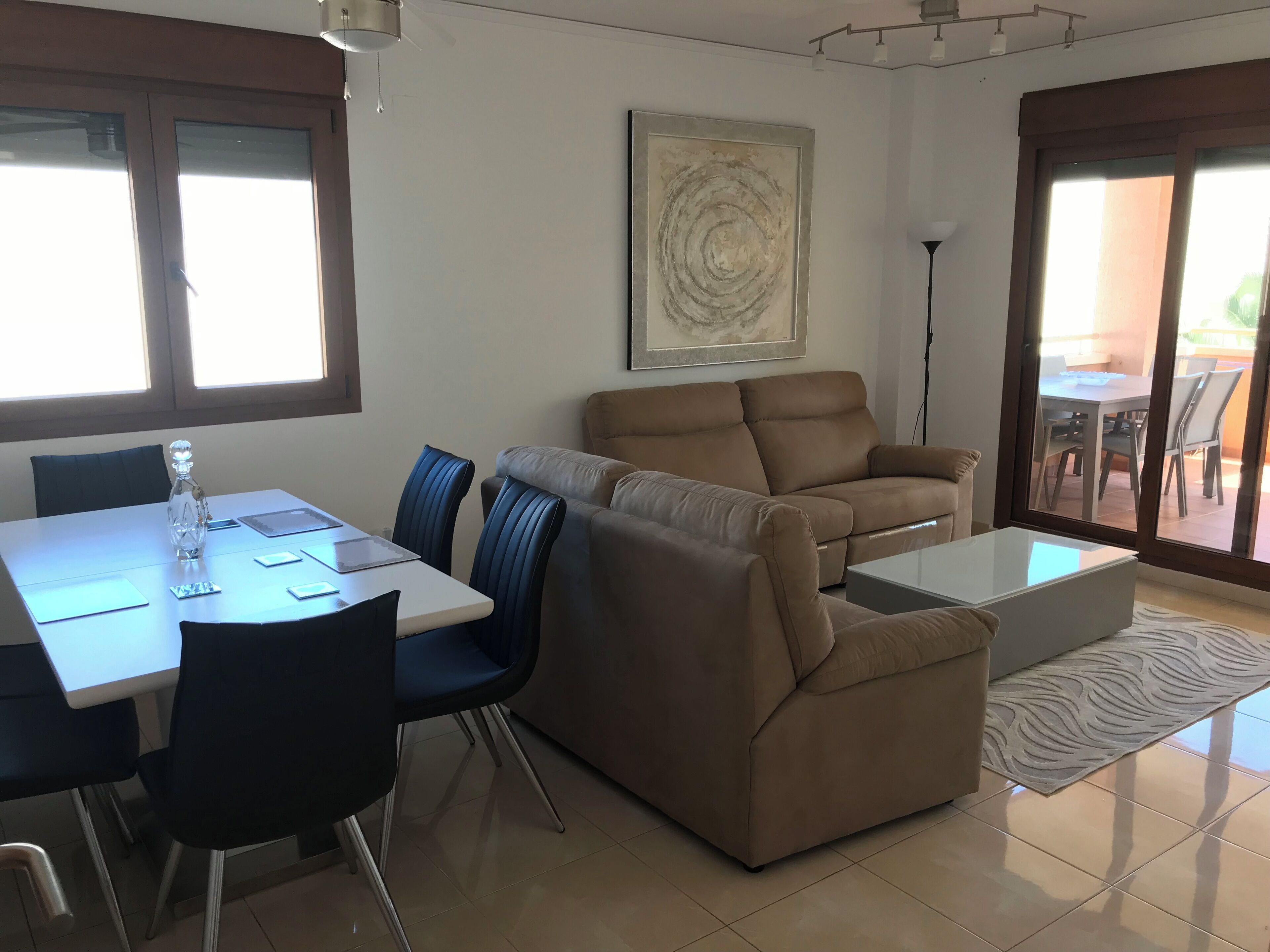 A living room with a sofa, a coffee table, and a dining table with chairs. There is a painting on the wall and a window with blinds.