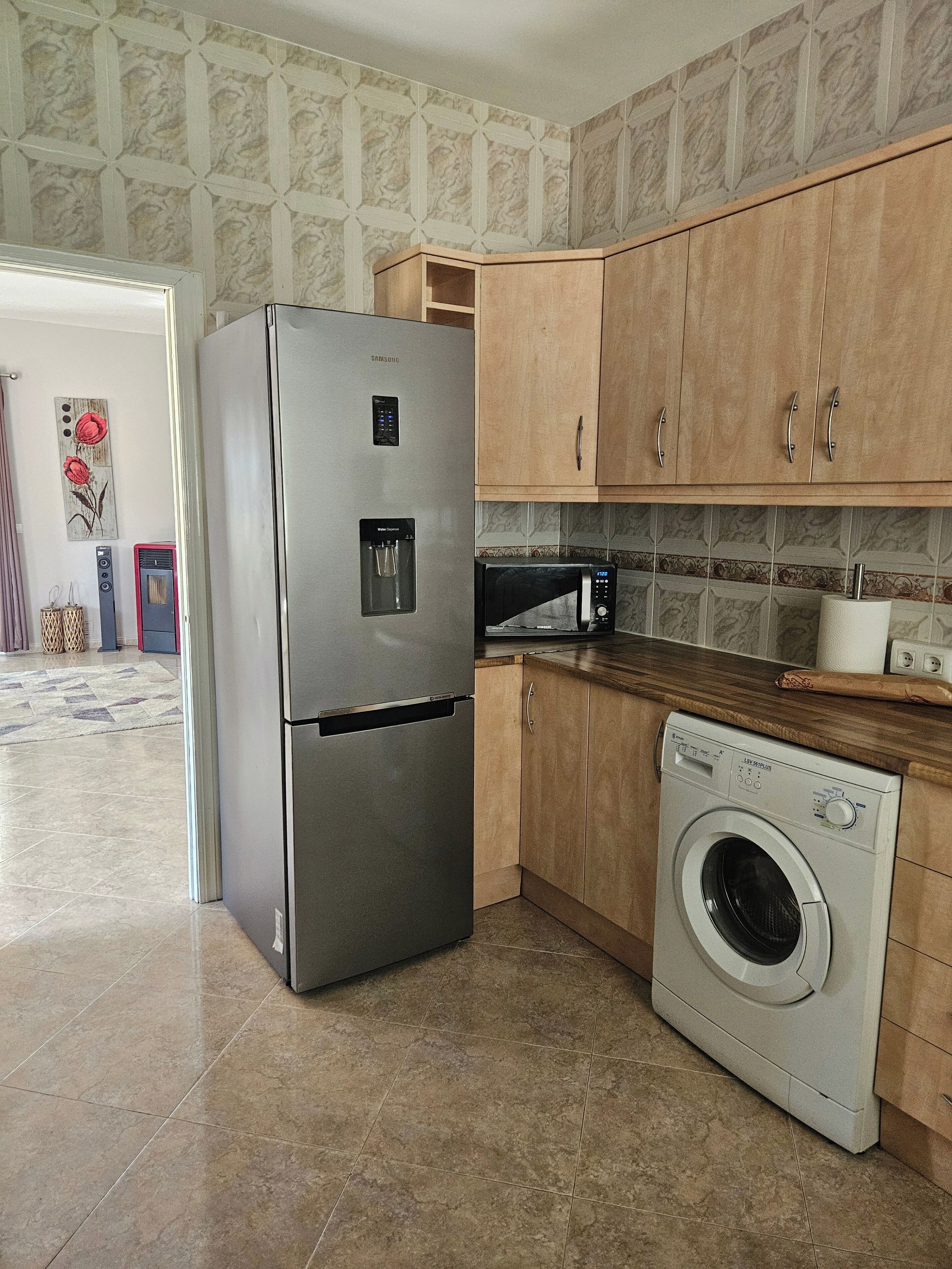Una cuina compacta amb nevera d'acer inoxidable, forn de microones integrat, armaris de fusta i rentadora de càrrega frontal.
