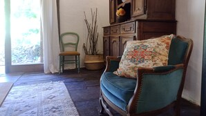 Een kamer met een houten schommelstoel, een groene stoel, een kast met lades en een raam met gordijnen.