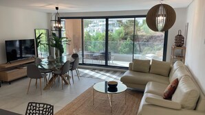 A modern living room with a TV, dining table, sofa, and large windows offering a view of the outdoors. - Modern Luxurious 3-bed Apartment in La Quinta Golf (Benahavis)