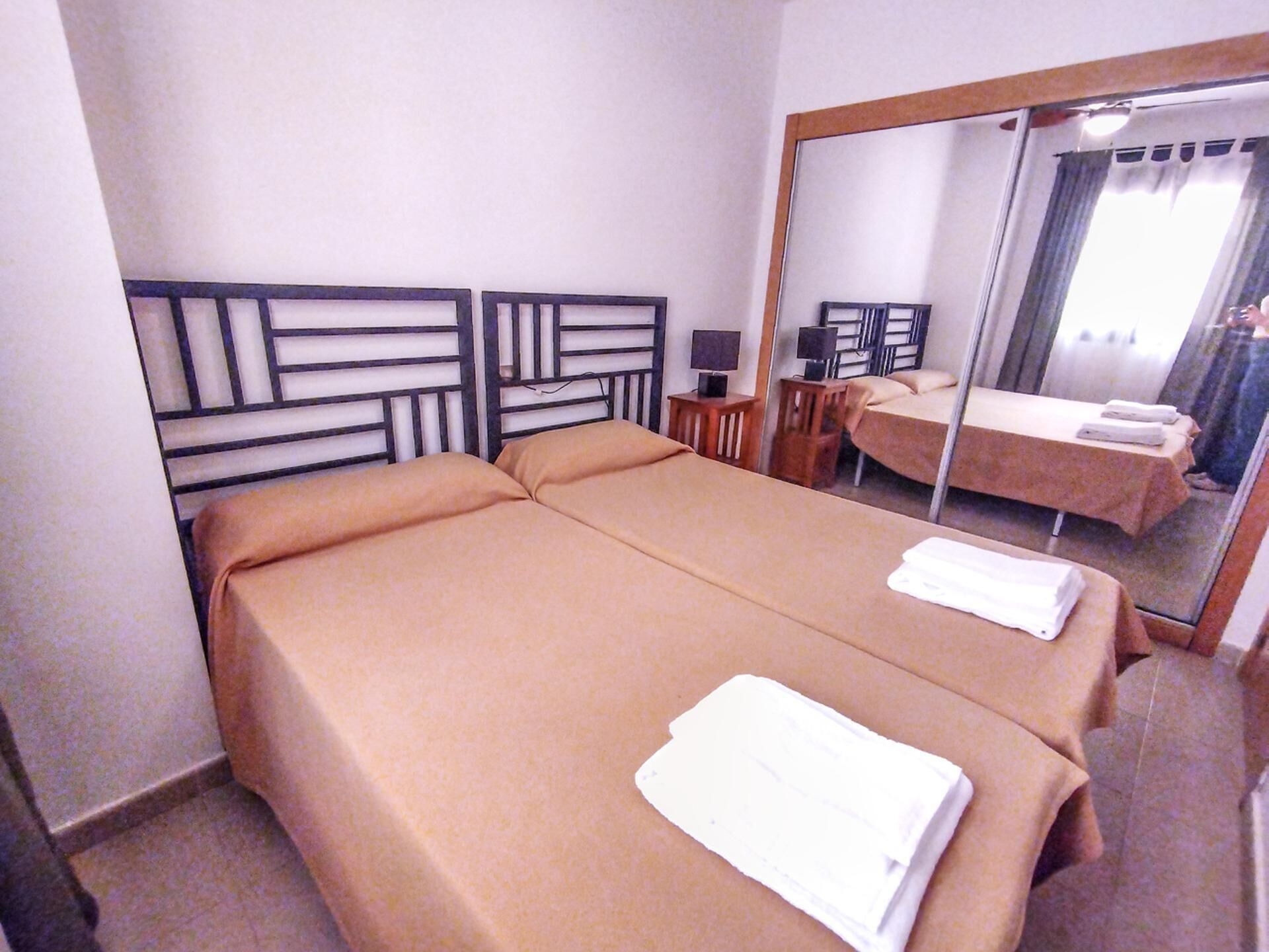 Ein Hotelzimmer mit Doppelbett, Nachttisch mit Lampe, Spiegel und Blick auf ein weiteres Zimmer mit Bett und Tisch.