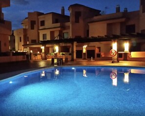 Une piscine à l’eau d’un bleu limpide, entourée de bâtiments dont l’éclairage est chaleureux.