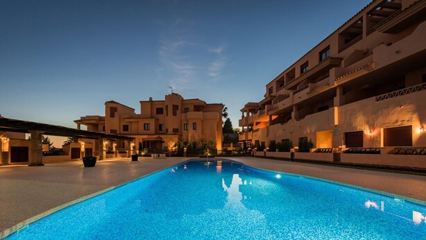 Una piscina con superficie de agua azul cristalina, rodeada de edificios con balcones e iluminación cálida.