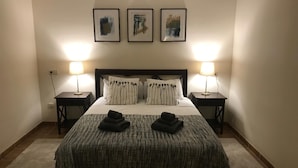 Una cama bien hecha con una colcha texturizada, dos almohadas y dos toallas dobladas. Hay dos mesitas de noche con lámparas, y tres obras de arte enmarcadas en la pared sobre la cama.