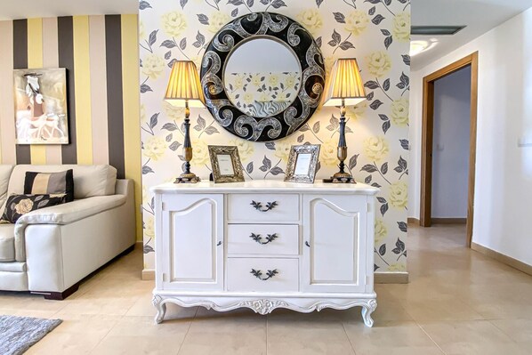 A living room with a white sofa, a white cabinet with decorative handles, two lamps, and a large mirror with a floral wallpaper background.