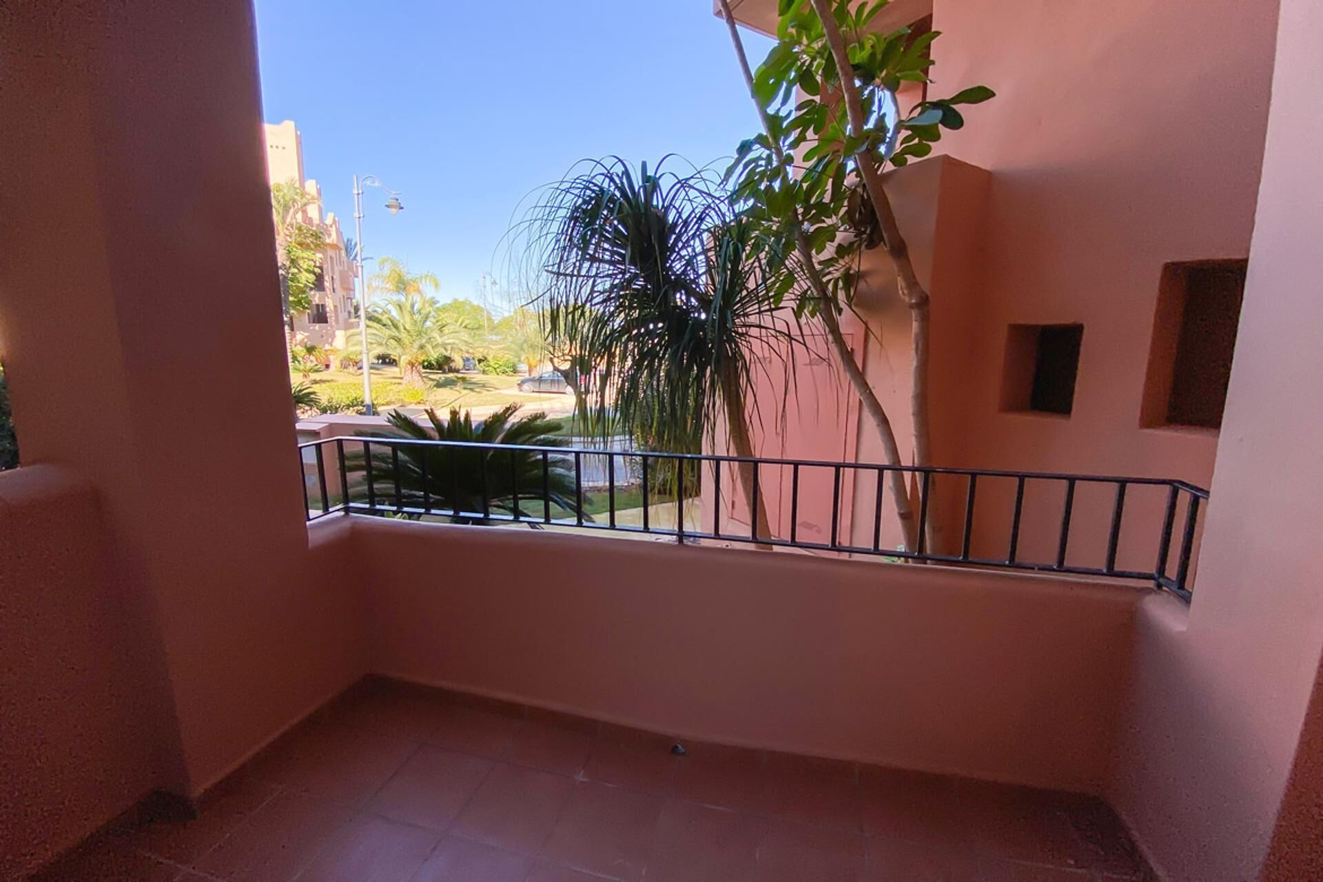 A balcony with a metal railing, a tiled floor, and a view of a residential area with palm trees and a building in the background.