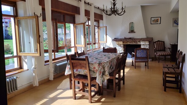 A dining area with a table set for four, surrounded by wooden chairs. There is a fireplace with a stone mantle, and a chandelier hangs from the ceiling. - Charming 6-bed Chalet in Llanes (Llanes)