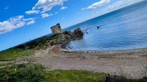 Ett kustlandskap med en förfallen stenstruktur nära vattenbrynet, en sandstrand och en klarblå himmel.